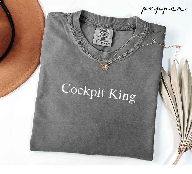 May include: A gray Comfort Colors t-shirt with the text "Cockpit King" in white. The shirt has a crew neck and short sleeves. A gold necklace with a pendant is draped on the shirt. The word "pepper" is written in cursive in the upper right corner.