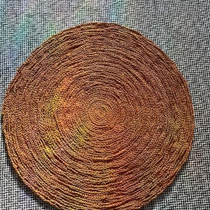 May include: A round, woven placemat in shades of brown and gold. The placemat is made of a textured material and has a spiral design. The background is a black and white patterned fabric.
