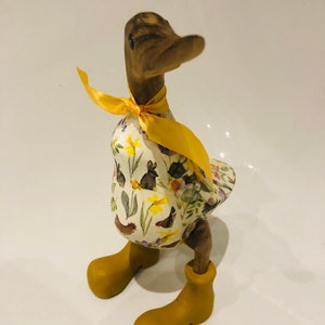 May include: A wooden duck figurine with a yellow bow and a floral print dress. The duck is wearing yellow boots and standing on a white surface.