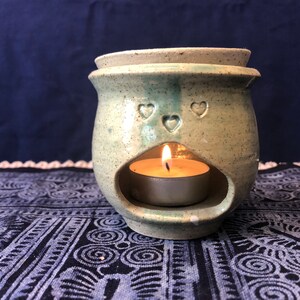 May include: A ceramic oil diffuser with a heart design and a teal and white glaze. A small tea light candle is burning inside the diffuser.