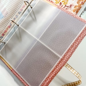 Puede incluir: Una carpeta de tres anillas con fundas de pl&aacute;stico transparente de cuatro bolsillos por p&aacute;gina. La cubierta es de tela con un estampado de lunares rosas y blancos, y lleva una cinta de cuadros vichy beige y blanca en el borde inferior.