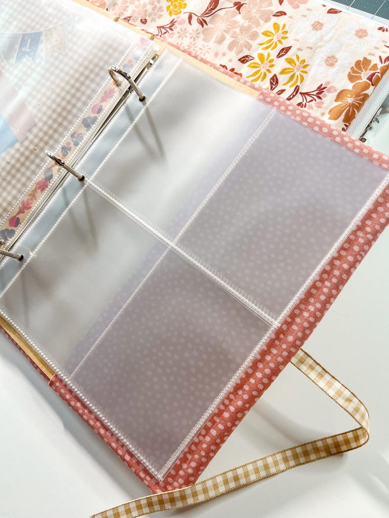 Puede incluir: Una carpeta de tres anillas con fundas de pl&aacute;stico transparente de cuatro bolsillos por p&aacute;gina. La cubierta es de tela con un estampado de lunares rosas y blancos, y lleva una cinta de cuadros vichy beige y blanca en el borde inferior.