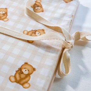 Puede incluir: Un coj&iacute;n rectangular de tela con un patr&oacute;n de cuadros vichy en beige claro y blanco, con un estampado repetitivo de ositos de peluche marrones. El coj&iacute;n incluye lazos de tela en espiga de color beige claro para sujetarlo.