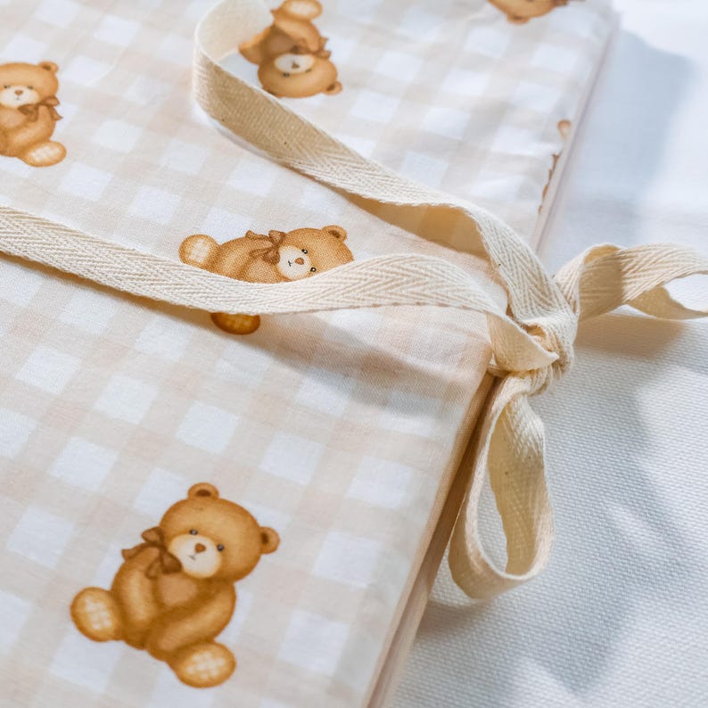 Puede incluir: Un coj&iacute;n rectangular de tela con un patr&oacute;n de cuadros vichy en beige claro y blanco, con un estampado repetitivo de ositos de peluche marrones. El coj&iacute;n incluye lazos de tela en espiga de color beige claro para sujetarlo.