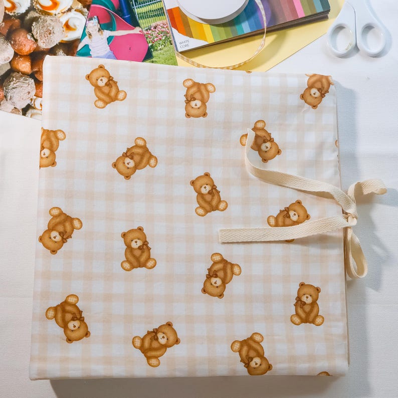Puede incluir: Una funda de tela cuadrada con un patr&oacute;n repetitivo de osos de peluche marrones sobre un fondo de cuadros vichy en beige claro y blanco. La funda incluye una cinta de tela de color crema unida al lado derecho para asegurar el cierre.