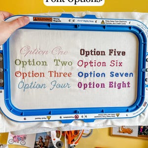 Puede incluir: Un bastidor de bordado azul sostiene una tela blanca con el texto Font Options en la parte superior. Ocho ejemplos de estilos de fuente numerados est&aacute;n bordados en la tela en varios colores, incluyendo Option One, Option Two, Option Three, Option Four, Option Five, Option Six, Option Seven y Option Eight.