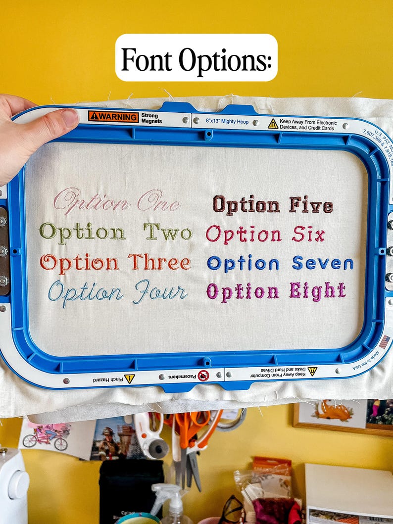 Puede incluir: Un bastidor de bordado azul sostiene una tela blanca con el texto Font Options en la parte superior. Ocho ejemplos de estilos de fuente numerados est&aacute;n bordados en la tela en varios colores, incluyendo Option One, Option Two, Option Three, Option Four, Option Five, Option Six, Option Seven y Option Eight.