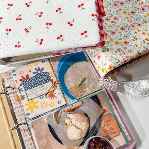 Puede incluir: Un archivador de recetas con una cubierta blanca con estampado de cerezas rojas y un lomo de cuadros rojos. Las p&aacute;ginas interiores muestran fotos de pan artesanal, un taz&oacute;n de sopa y el texto Happy Hearts Homemade Artisan Bread Yum! y pinch of this, dash of that.