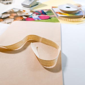 Puede incluir: Una tira de cinta de sarga de algod&oacute;n beige con patr&oacute;n de espiga sobre una superficie lisa de color marr&oacute;n claro. La cinta de tela tejida presenta un patr&oacute;n en forma de V en toda su textura, con un extremo formando un bucle sobre el material.