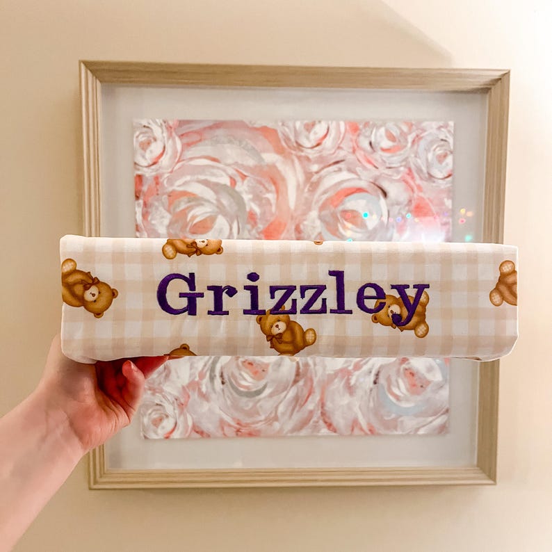 Puede incluir: Un art&iacute;culo de tela rectangular con un patr&oacute;n de cuadros vichy en beige claro y blanco, con un estampado repetitivo de peque&ntilde;os osos de peluche marrones. El nombre "Grizzley" est&aacute; bordado en letras may&uacute;sculas moradas, negritas y sin remates.