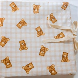 Puede incluir: Un art&iacute;culo de tela con un patr&oacute;n repetitivo de osos de peluche marrones sobre un fondo de cuadros vichy en beige claro y blanco. El textil incluye una cinta de espiga color crema unida al lateral para asegurar el material plegado.