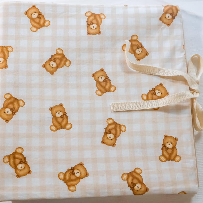 Puede incluir: Un art&iacute;culo de tela con un patr&oacute;n repetitivo de osos de peluche marrones sobre un fondo de cuadros vichy en beige claro y blanco. El textil incluye una cinta de espiga color crema unida al lateral para asegurar el material plegado.