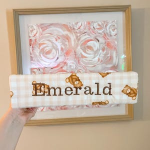 Puede incluir: Un objeto rectangular de tela acolchada con un patr&oacute;n de cuadros vichy en beige claro y blanco, decorado con peque&ntilde;as ilustraciones de ositos de peluche marrones. El nombre Emerald est&aacute; bordado en hilo marr&oacute;n oscuro en el centro.