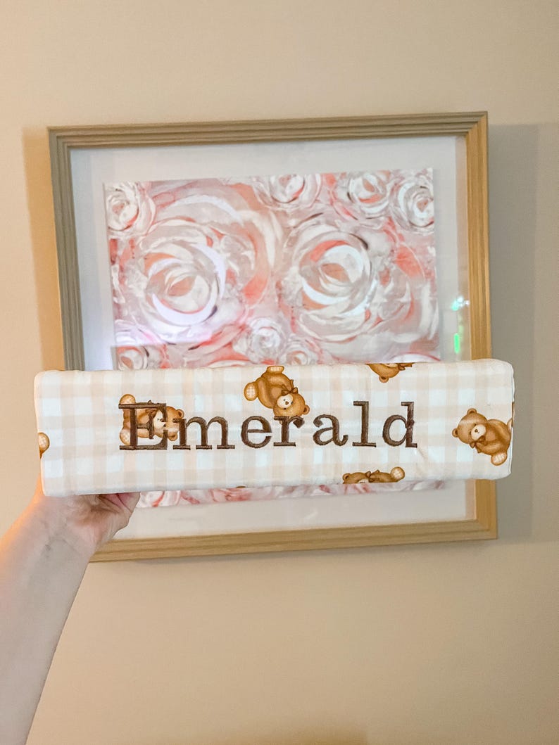 Puede incluir: Un objeto rectangular de tela acolchada con un patr&oacute;n de cuadros vichy en beige claro y blanco, decorado con peque&ntilde;as ilustraciones de ositos de peluche marrones. El nombre Emerald est&aacute; bordado en hilo marr&oacute;n oscuro en el centro.