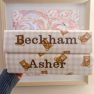 Puede incluir: Dos art&iacute;culos rectangulares apilados con un patr&oacute;n de cuadros vichy en beige claro y blanco. Cada art&iacute;culo muestra un nombre bordado en marr&oacute;n, Beckham y Asher, rodeado de peque&ntilde;as ilustraciones de ositos marrones sobre la tela.