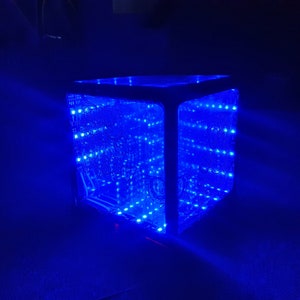 Può includere: Un cubo infinito blu brillante con numerose piccole luci a LED. L'interno del cubo riflette le luci, creando un effetto affascinante. Il cubo è posto su uno sfondo scuro, esaltando la vibrante illuminazione blu.