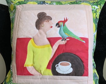 May include: Square decorative cushion with a painted design. The cushion features a woman in a yellow dress with a parrot. The background is red, with a cup of coffee. The border has a lemon and leaf pattern.