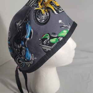 May include: A gray surgical scrub cap featuring colorful motorcycle graphics. The cap has a black trim and black ties for securing. Motorcycles in yellow, blue, and green are printed on the fabric.
