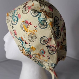 May include: A cream-colored surgical scrub cap featuring a repeating pattern of colorful bicycles with flower baskets. The bicycles are in shades of yellow, blue, and pink. The cap has ties at the back for an adjustable fit.