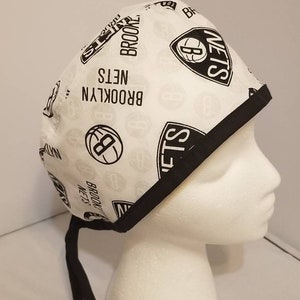 May include: White surgical scrub hat with black elastic ties. The hat has a repeating pattern of the Brooklyn Nets logo, the team's name, and a basketball.