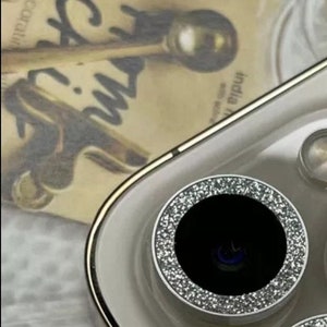 May include: A close-up of a silver glitter phone camera lens cover. The cover is round and has a black center with a small, round, blue lens.