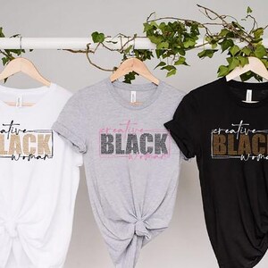 May include: Three t-shirts in white, gray, and black hang on wooden hangers. Each shirt features the text "creative BLACK woman" in a rectangular design. The text is in different colors, including gold, pink, and white.