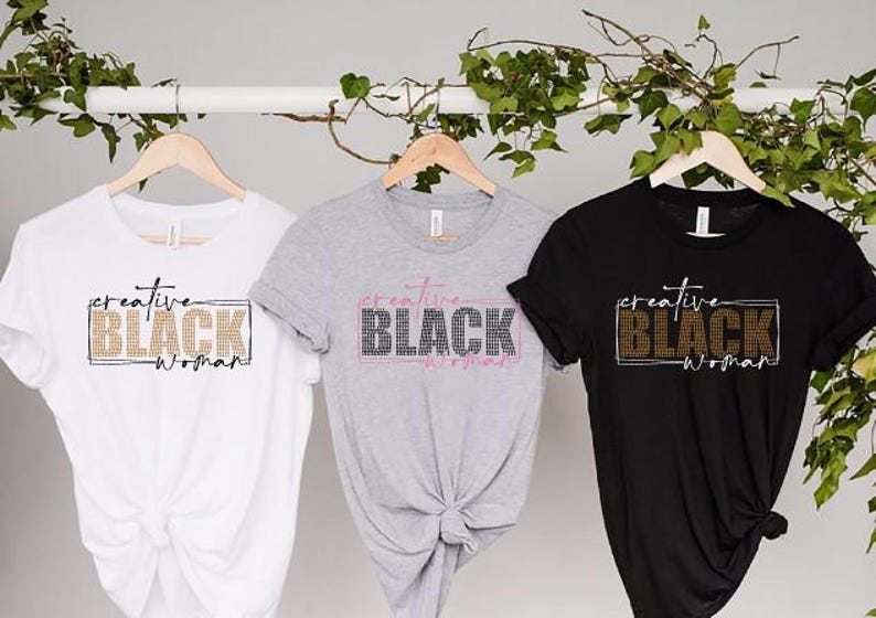 May include: Three t-shirts in white, gray, and black hang on wooden hangers. Each shirt features the text "creative BLACK woman" in a rectangular design. The text is in different colors, including gold, pink, and white.