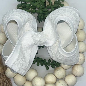 May include: A large, white hair bow with a textured lace overlay and a satin ribbon base. The bow is centered on a wreath of white wooden beads, with green faux succulents in the background. A natural fiber tassel is visible on the left.