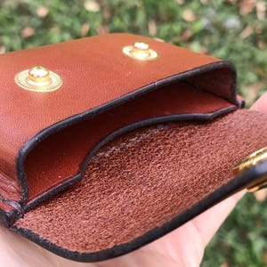 May include: A brown leather pouch featuring a smooth exterior and a textured interior. The pouch is secured with two gold-toned metal snap buttons on the front flap, which is designed to fold over the main compartment opening.