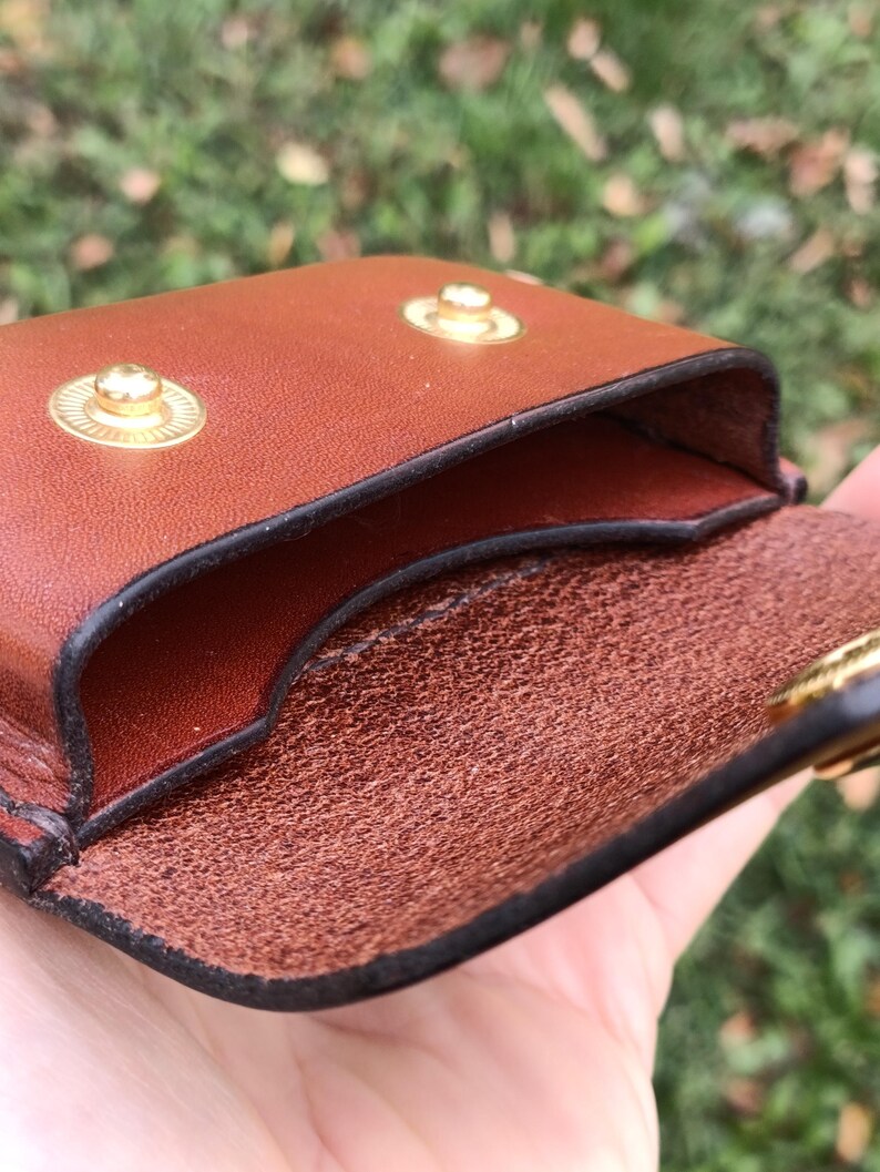 May include: A brown leather pouch featuring a smooth exterior and a textured interior. The pouch is secured with two gold-toned metal snap buttons on the front flap, which is designed to fold over the main compartment opening.