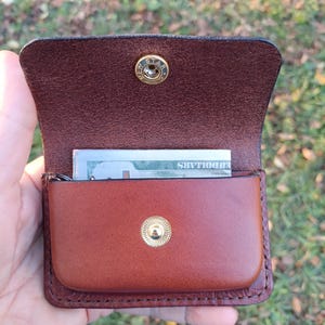 May include: A brown leather wallet with a flap closure featuring a gold-coloured metal snap button. The interior contains a folded US dollar bill. The wallet has visible stitching along the edges and a textured finish on the inner flap.