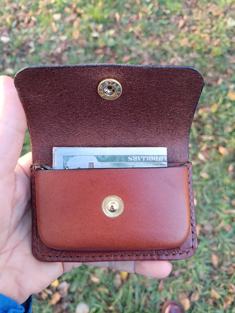 May include: A brown leather wallet with a flap closure featuring a gold-coloured metal snap button. The interior contains a folded US dollar bill. The wallet has visible stitching along the edges and a textured finish on the inner flap.