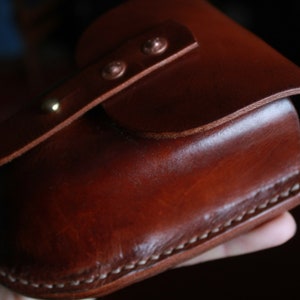 May include: A handcrafted, brown leather case with a curved top and a flap secured by two rivets. The case features visible white stitching along the bottom edge and a smooth, polished finish. The leather has a rich, warm tone.