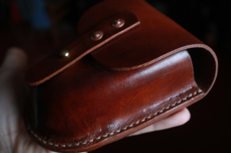 May include: A handcrafted, brown leather case with a curved top and a flap secured by two rivets. The case features visible white stitching along the bottom edge and a smooth, polished finish. The leather has a rich, warm tone.