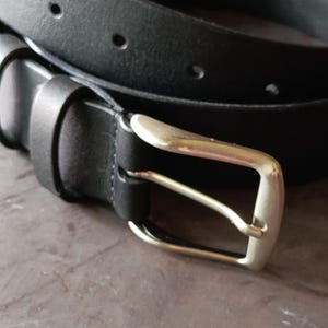 Puede incluir: Un cintur&oacute;n negro con un acabado mate liso y una hebilla de metal plateado. La correa del cintur&oacute;n incluye una serie de orificios circulares espaciados uniformemente para el ajuste y una trabilla de cuero negro a juego para asegurar el extremo.
