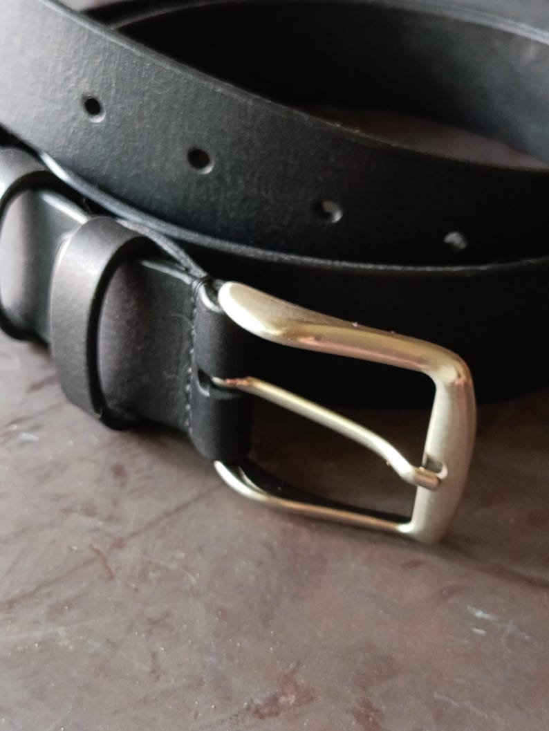 Puede incluir: Un cintur&oacute;n negro con un acabado mate liso y una hebilla de metal plateado. La correa del cintur&oacute;n incluye una serie de orificios circulares espaciados uniformemente para el ajuste y una trabilla de cuero negro a juego para asegurar el extremo.