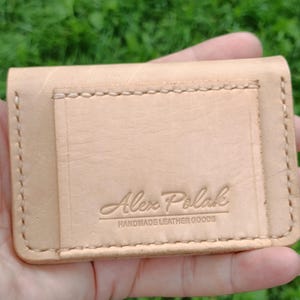 May include: A light tan leather card holder featuring visible stitching along the edges and a rectangular pocket on the front. The leather is embossed with the text Alex Polak HANDMADE LEATHER GOODS in a clear, centered font on the front pocket.