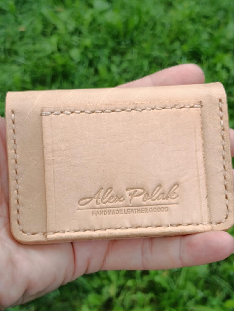 May include: A light tan leather card holder featuring visible stitching along the edges and a rectangular pocket on the front. The leather is embossed with the text Alex Polak HANDMADE LEATHER GOODS in a clear, centered font on the front pocket.