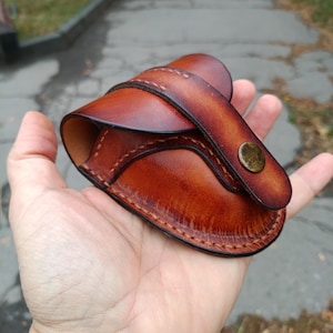K&ouml;nnte beinhalten: Eine handgefertigte braune Leder-Tasche mit geschwungener Form, gen&auml;hter Kante und einer Klappe, die mit einem Messingknopf befestigt ist. Die Tasche wird in einer Hand gehalten, wodurch die Farbe und Textur des Leders hervorgehoben werden.