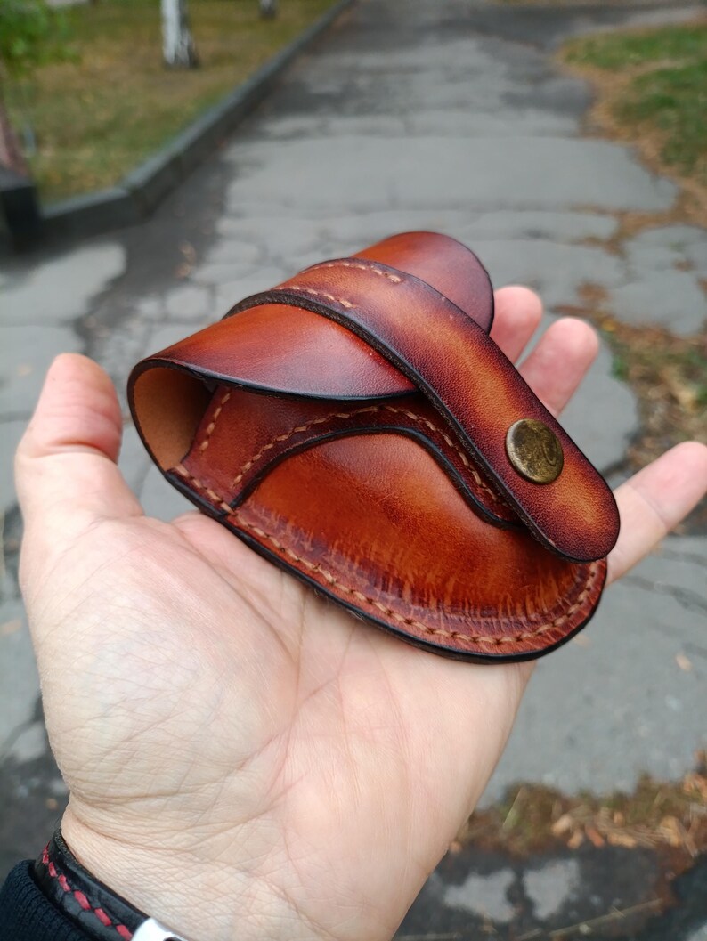 K&ouml;nnte beinhalten: Eine handgefertigte braune Leder-Tasche mit geschwungener Form, gen&auml;hter Kante und einer Klappe, die mit einem Messingknopf befestigt ist. Die Tasche wird in einer Hand gehalten, wodurch die Farbe und Textur des Leders hervorgehoben werden.