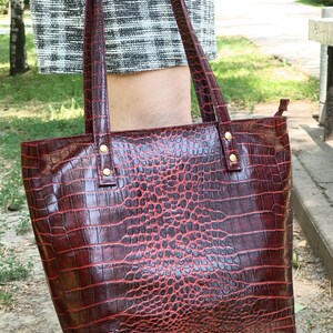 May include: A deep red tote bag with a glossy crocodile skin texture pattern. The bag features two vertical shoulder straps attached with gold-toned metal rivets at the top. The surface has a dark, embossed scale design throughout the exterior.