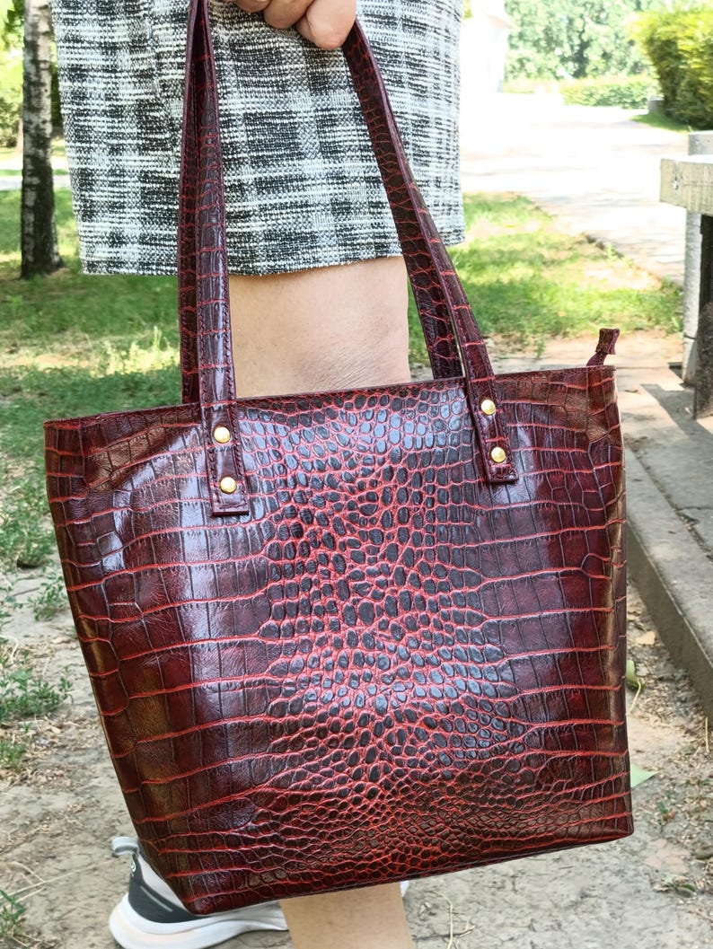 May include: A deep red tote bag with a glossy crocodile skin texture pattern. The bag features two vertical shoulder straps attached with gold-toned metal rivets at the top. The surface has a dark, embossed scale design throughout the exterior.