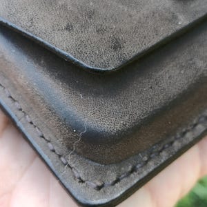 May include: A dark charcoal gray wallet with a textured surface and visible stitching along the edges. The wallet features a flap closure secured by a single, smooth, circular brass-toned metal snap button centered on the upper portion.