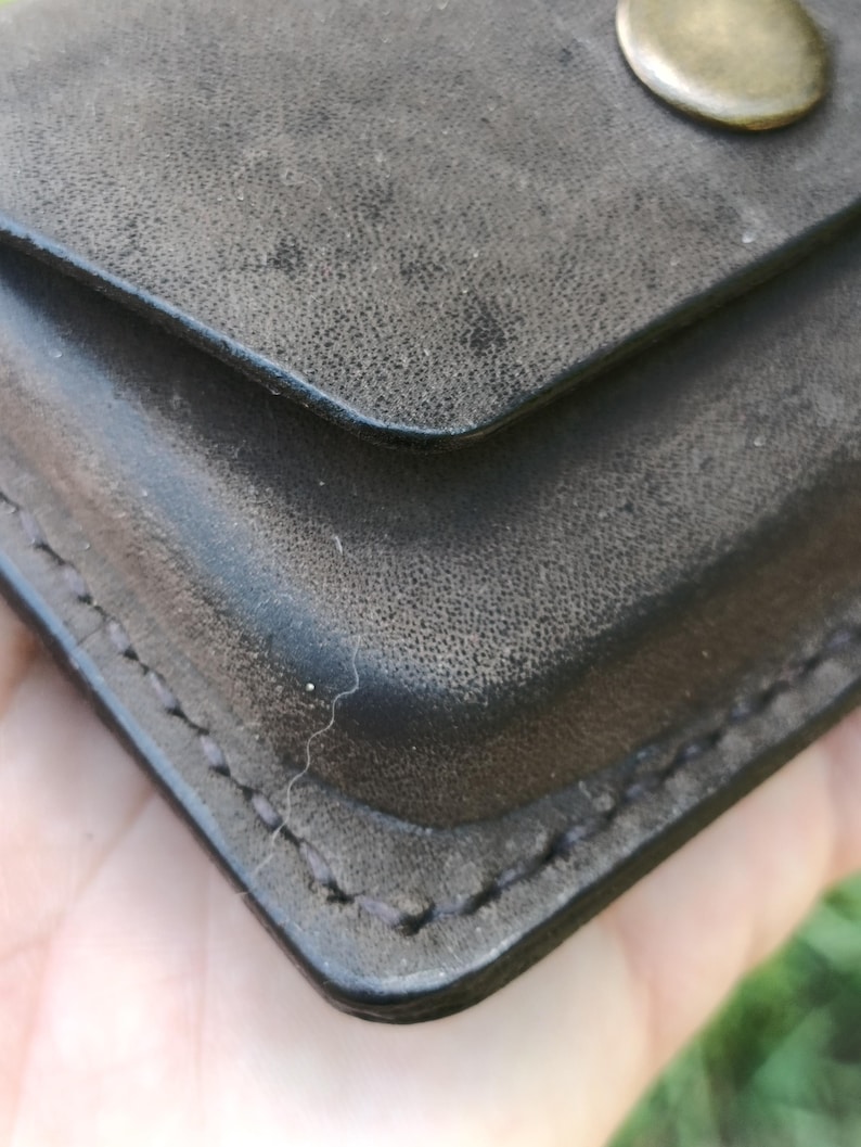 May include: A dark charcoal gray wallet with a textured surface and visible stitching along the edges. The wallet features a flap closure secured by a single, smooth, circular brass-toned metal snap button centered on the upper portion.