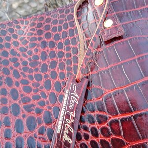 May include: A deep red tote bag with a glossy crocodile skin texture. The bag features a matching rectangular leather tag hanging from a thin cord, embossed with the text Alex Pohl Handmade Leather Goods and two gold-toned metal rivets.