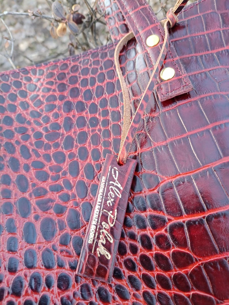 May include: A deep red tote bag with a glossy crocodile skin texture. The bag features a matching rectangular leather tag hanging from a thin cord, embossed with the text Alex Pohl Handmade Leather Goods and two gold-toned metal rivets.