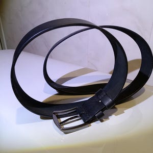 May include: A black leather belt with a silver buckle. The belt is looped on a white surface. The buckle is rectangular with a single prong.