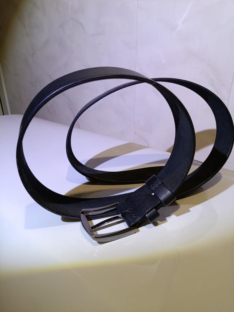 May include: A black leather belt with a silver buckle. The belt is looped on a white surface. The buckle is rectangular with a single prong.
