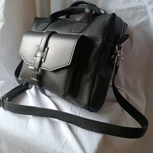 May include: Black leather messenger bag with a flap pocket, top handle, and adjustable shoulder strap. The bag has a textured finish and silver-tone hardware. The bag is designed with a front flap pocket secured by a buckle closure.