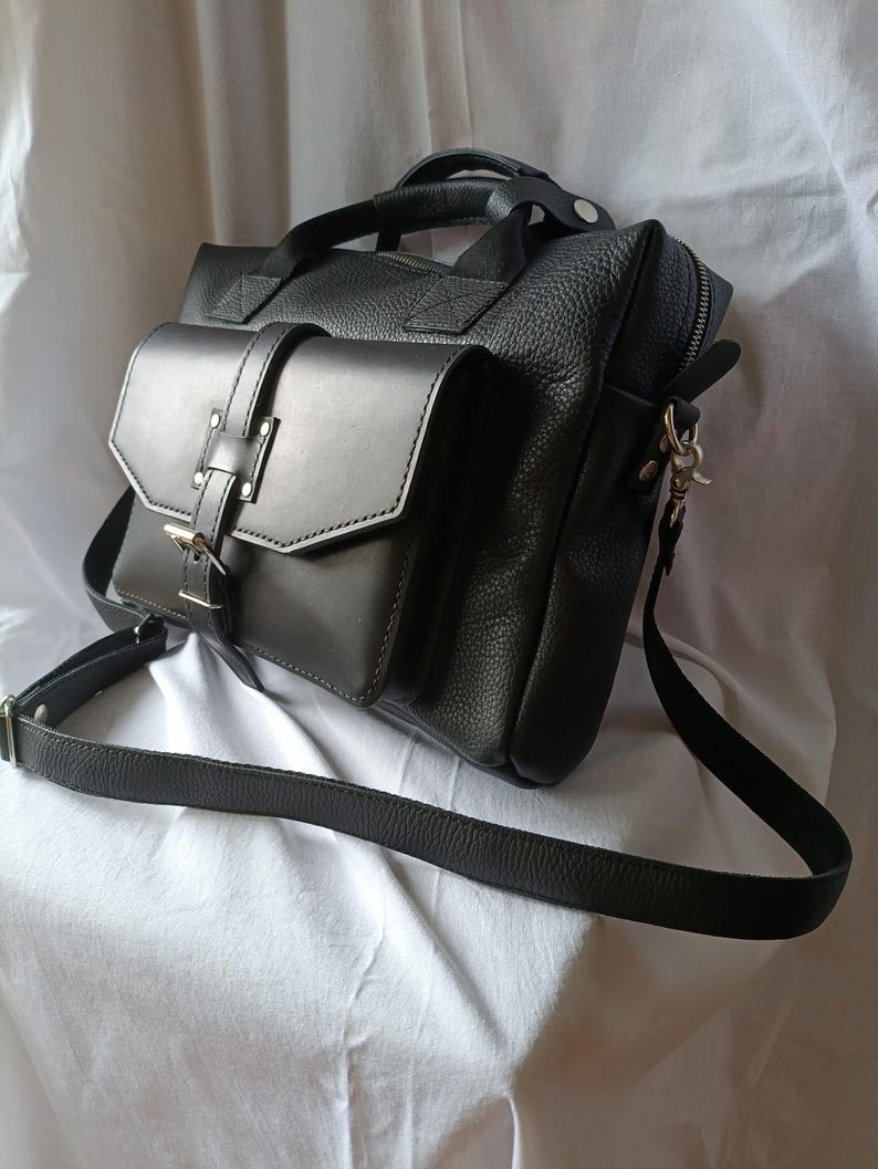 Puede incluir: Bolso mensajero de cuero negro con un bolsillo con solapa, asa superior y correa de hombro ajustable. El bolso tiene un acabado texturizado y herrajes plateados. El bolso est&aacute; dise&ntilde;ado con un bolsillo frontal con solapa asegurado con un cierre de hebilla.
